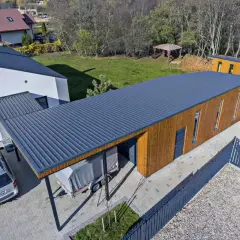 Nowoczesny budynek z drewnianą elewacją i ciemnym, **dachem jednospadowym**. Obok stoi samochód i przyczepa pod zadaszeniem.