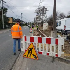 Robotnicy w kamizelkach odblaskowych pracują na budowie przy drodze, ogrodzonej barierkami.