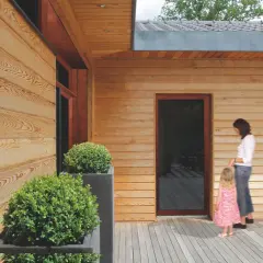 Drewniana elewacja domu, zabezpieczona impregnatem, z kobietą i dzieckiem na tarasie.