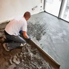 Budowlaniec klęczy, wyrównując świeży jastrych metalową listwą. Prace budowlane w nowym mieszkaniu.