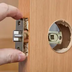 Dłoń montuje mechanizm zamka w drewnianych drzwiach. Dowiedz się, ile kosztuje wymiana zamka w drzwiach.