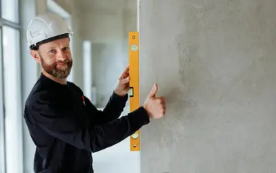 Budowlaniec w kasku sprawdza pionową ścianę za pomocą poziomicy. Kciuk w górę potwierdza, że wszystko jest idealnie.