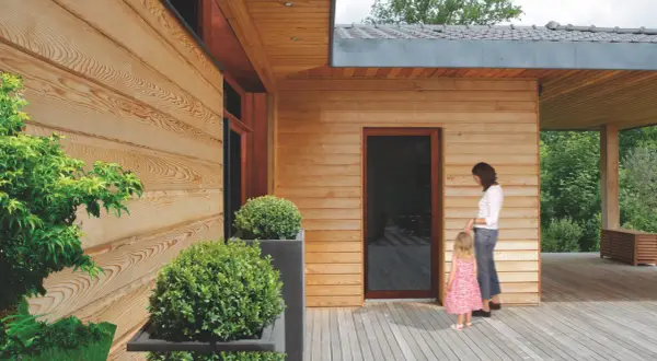 Drewniana elewacja domu, zabezpieczona impregnatem, z kobietą i dzieckiem na tarasie.