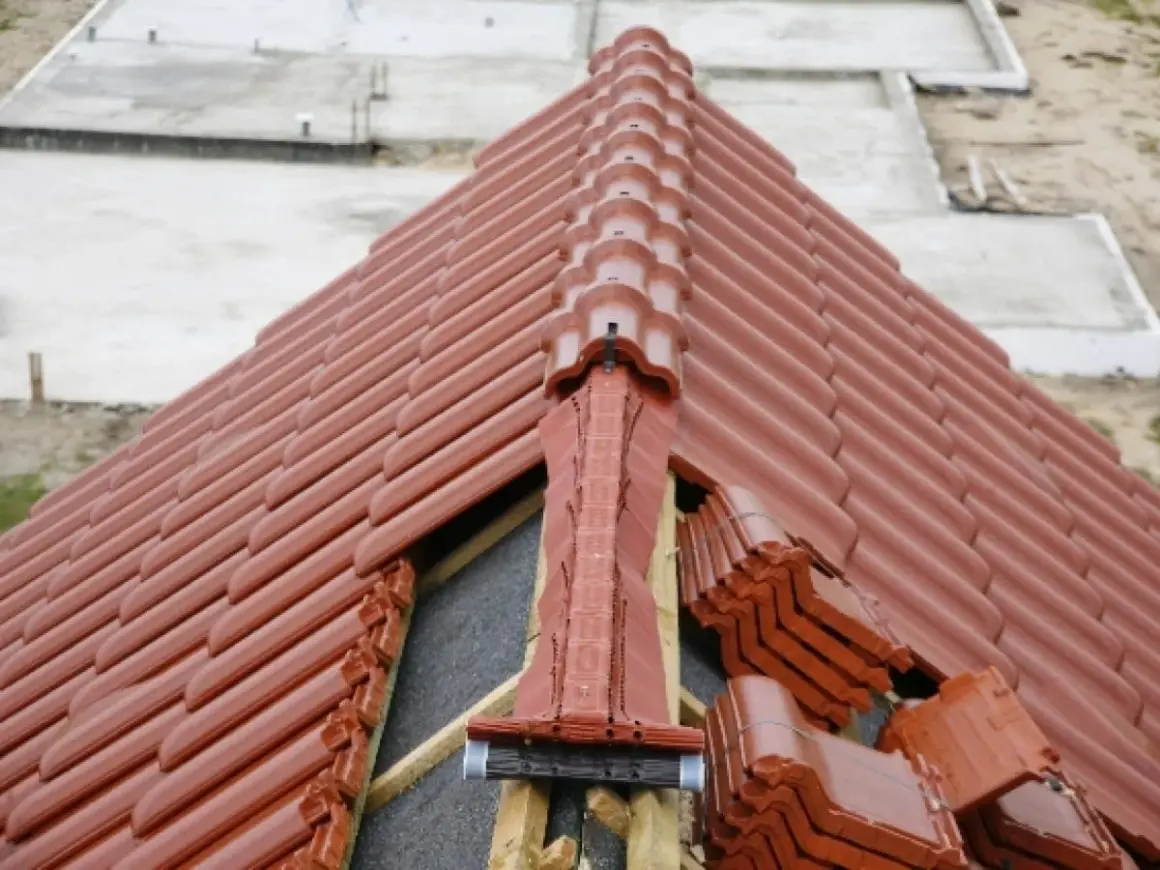 Nowy dach z brązowej dachówki. Widoczne miejsce przecięcia dwóch połaci dachowych, z elementami wentylacyjnymi i zapasem dachówek.