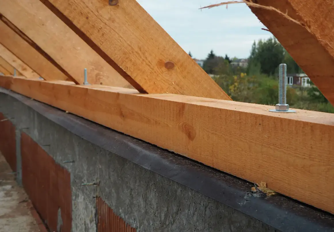 Drewniana murłata na betonowym wieńcu, przygotowana do montażu więźby dachowej. Widać śruby mocujące.