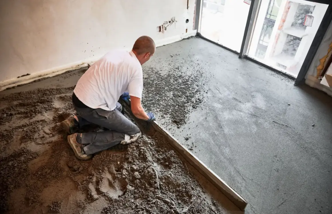 Budowlaniec klęczy, wyrównując świeży jastrych metalową listwą. Prace budowlane w nowym mieszkaniu.