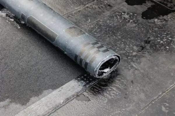 Zrolowana papa na zimno, z widocznymi czarnymi pasami i kratką, leży na mokrej, ciemnej powierzchni.