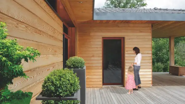 Drewniana elewacja domu, zabezpieczona impregnatem, z kobietą i dzieckiem na tarasie.