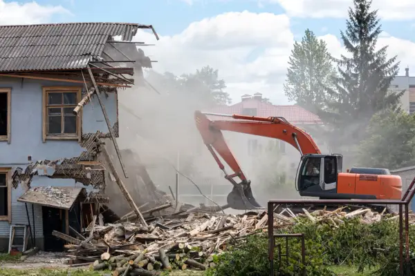Pomarańczowy koparka prowadzi rozbiórkę budynku, wzbijając tumany kurzu. Ruiny i drewno leżą na ziemi.